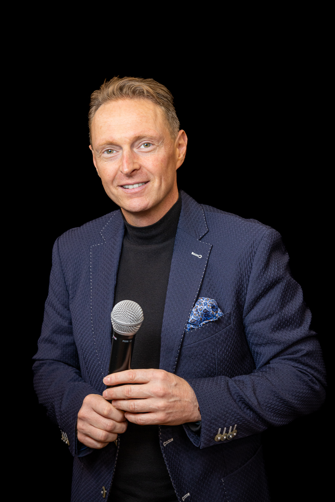 Franz Wulz, der einen blau gemusterten Blazer und einen schwarzen Rollkragenpullover trägt, hält ein Mikrofon und lächelt in die Kamera vor einem schwarzen Hintergrund.